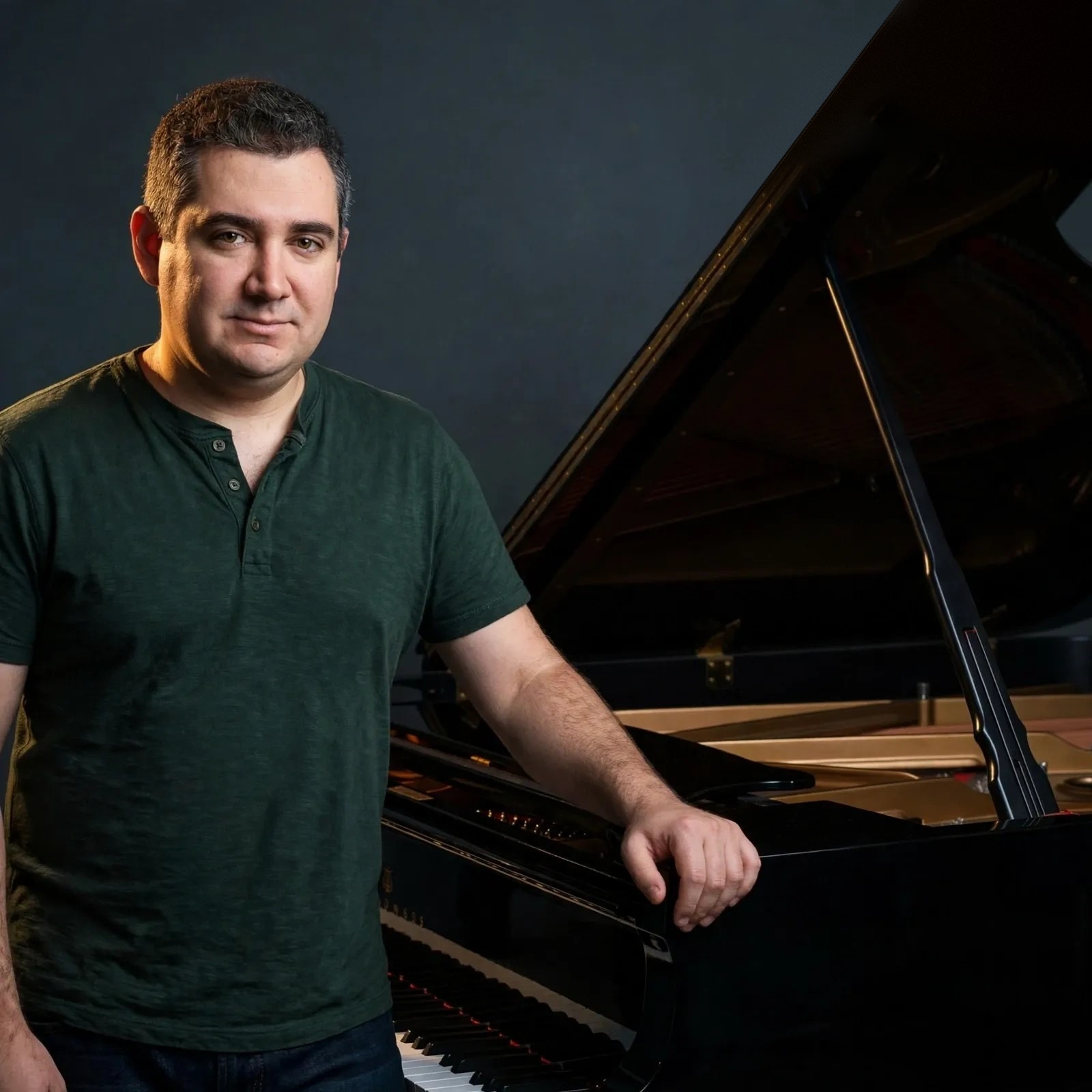 Jason Solowsky in a green henley standing beside a black grand piano with its lid raised, his hand resting on the piano