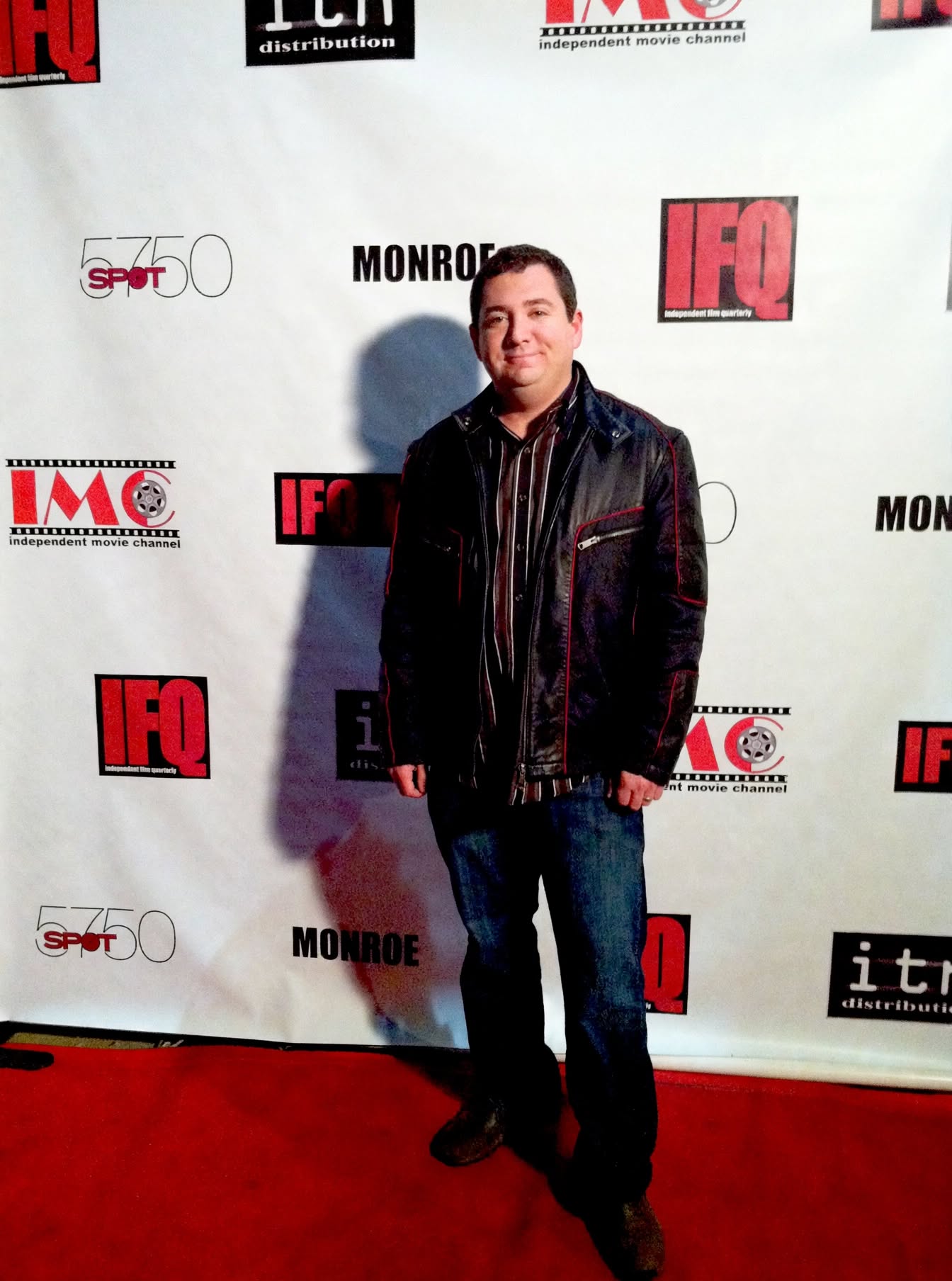Jason Solowsky in a black leather jacket standing on a red carpet in front of a step-and-repeat backdrop