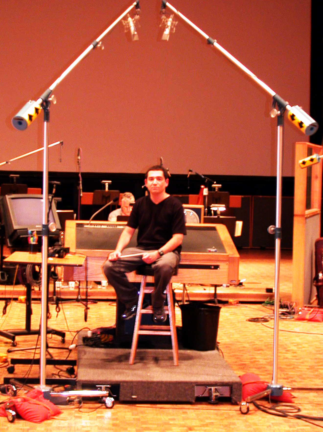 Jason Solowsky seated on the conductor's podium on an empty scoring stage, holding a baton, with microphone booms overhead
